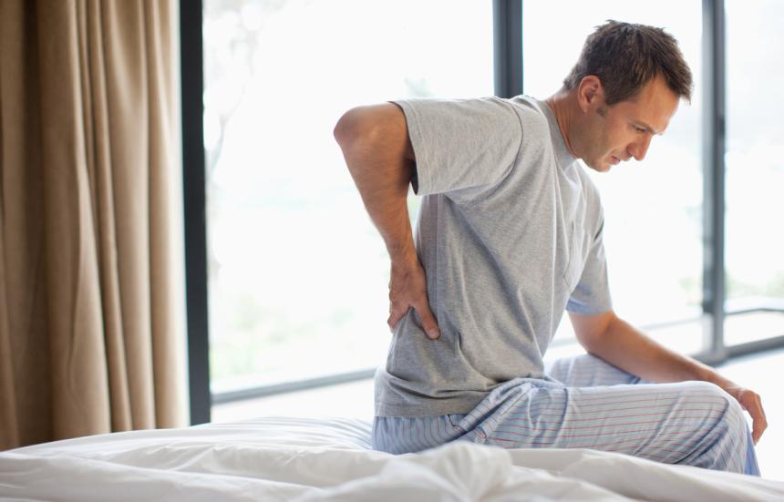 Middle aged man sitting on the edge of his bed, gripping his lower back.